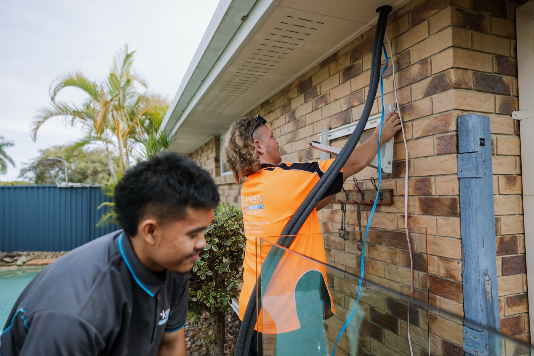 Parker Air technicians routing refrigerant pipe along brick exterior wall near pool